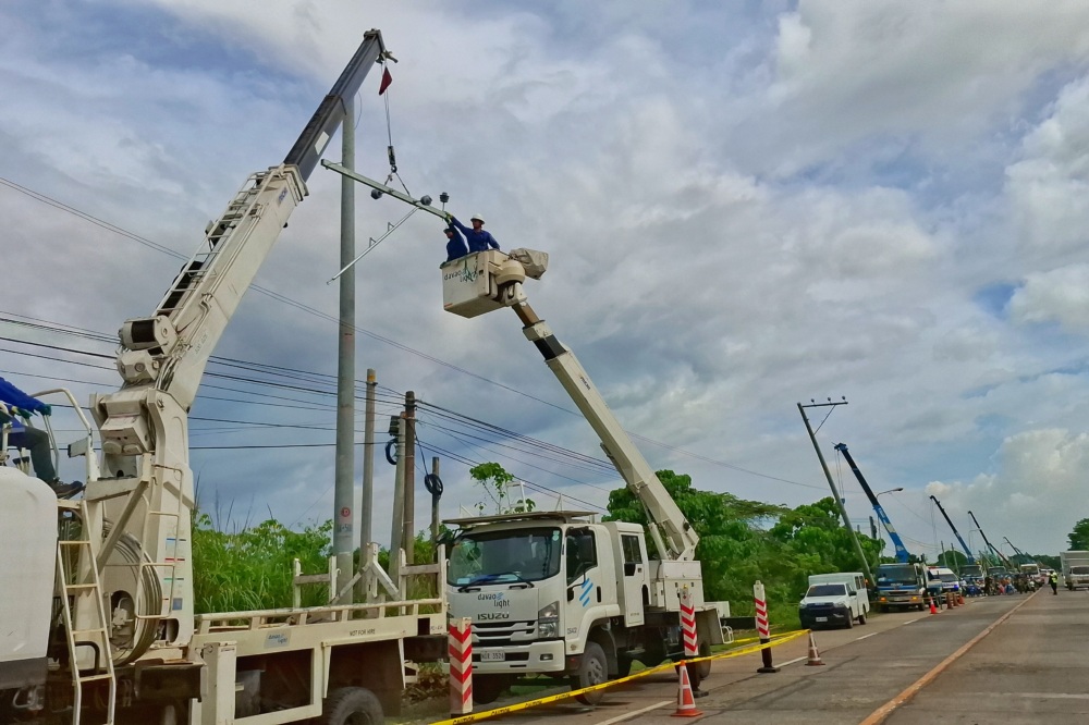 Men in a bucket attached to a crane attached to a truck work on constructing a power line on the side of a road.