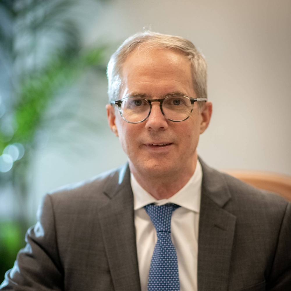 Head shot photo of a white man with glasses and a blue tie in front of a soft focus background.