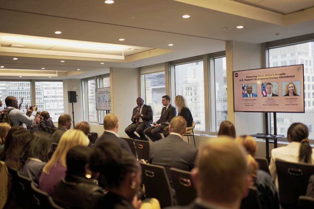 Photo of a panel discussion between three people sitting on stools in front of a full room in front of a set of windows.